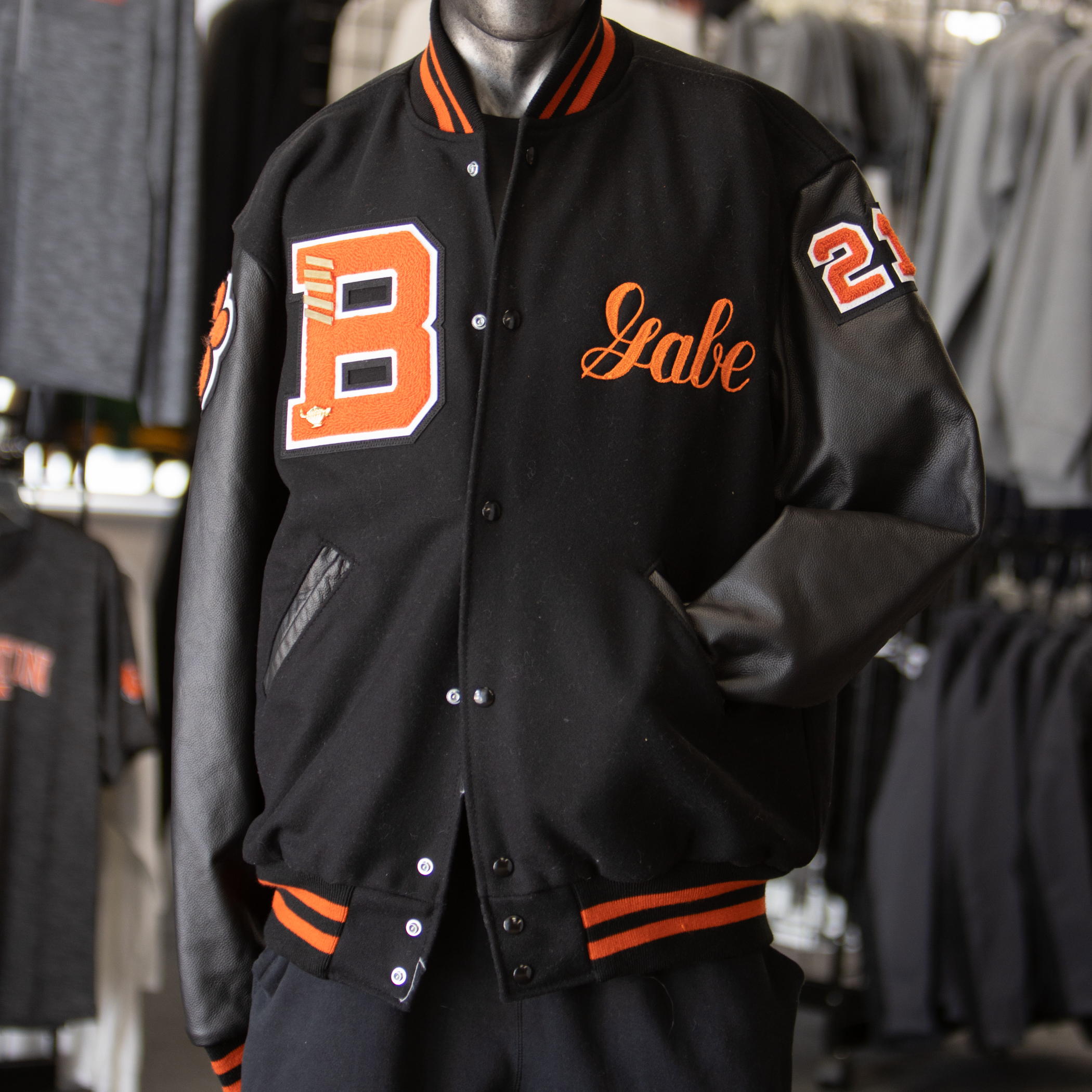 A black varsity jacket with orange and white details is displayed on a mannequin. The body of the jacket is black wool, and the sleeves look like black leather. There’s a big orange letter B patch on the left chest, and the name “Gabe” stitched in orange cursive on the right chest. The left sleeve has the number 21 in orange. The collar, cuffs, and waistband all have black-and-orange stripes. The mannequin is standing in a clothing store surrounded by racks of shirts.