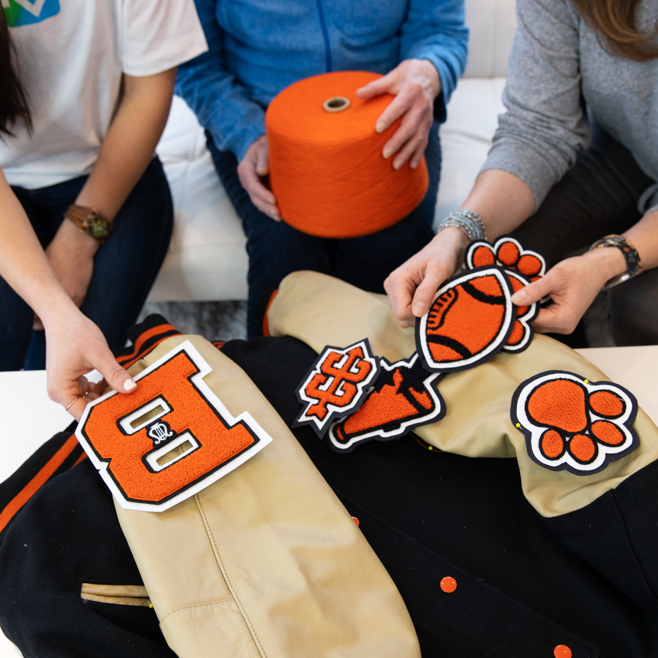 Three people are sitting around a table, placing bright orange-and-black sports patches onto a varsity jacket. One person is holding a big cone of orange yarn. The patches include a large letter B, a paw print, a football, and a megaphone. The jacket they’re decorating has light tan sleeves and a black body. The scene looks like a custom patch-design session.
